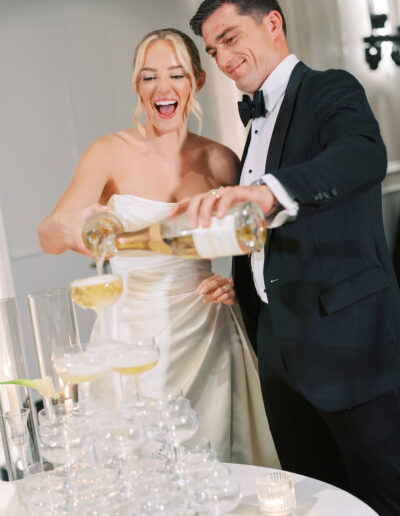 A bride and groom in formal attire pour champagne together into a tiered tower of glasses at a wedding reception.