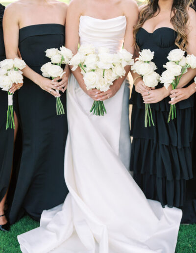 Three women in formal dresses holding white rose bouquets; the woman in the center wears a white gown, while the others wear black dresses.