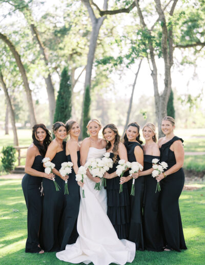A bride in a white dress stands outdoors with seven bridesmaids in black dresses, all holding white bouquets, with trees and grass in the background.