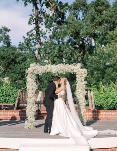 A bride and groom kiss under a white floral arch during an outdoor wedding ceremony, surrounded by greenery and trees.