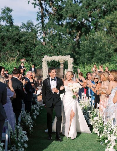 A bride and groom walk down an outdoor aisle lined with white flowers as guests stand and applaud.