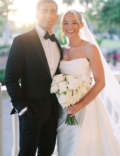A bride in a white gown holds a bouquet of white roses and stands next to a groom in a black tuxedo, both smiling at the camera outdoors.