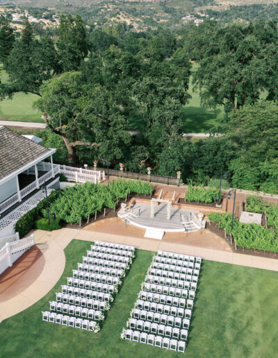 Rows of white chairs are arranged on a lawn facing a small outdoor stage, with trees and hills in the background.