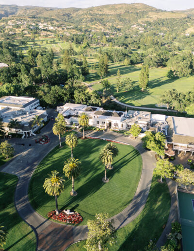 Aerial view of a circular driveway with palm trees, surrounded by buildings and a golf course in a hilly, green landscape.