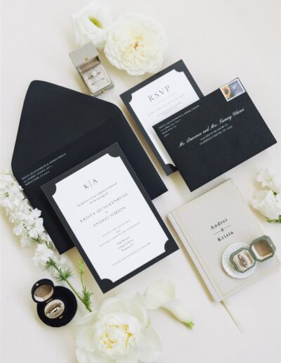 A flat lay of black and white wedding invitations, RSVP card, envelope, wedding rings in boxes, a vow booklet, and white flowers arranged on a light background.
