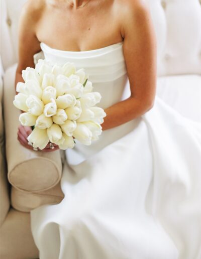 A woman in a white strapless dress sits on a beige couch, holding a bouquet of white tulips in her lap.