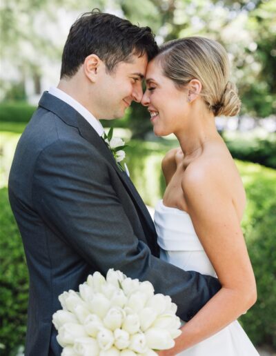 A bride and groom stand close together outdoors, smiling with foreheads touching. The bride holds a bouquet of white flowers.