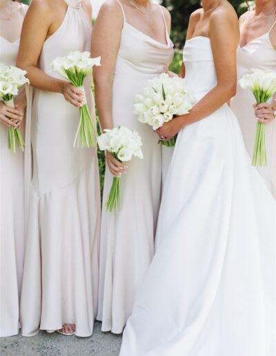 Four women in light-colored dresses, one in a white wedding gown, each holding a bouquet of white flowers, standing side by side.