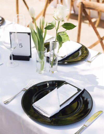 A table setting with a black plate, folded white napkin, menu card, and glass vases holding white tulips and a rose on a white tablecloth.