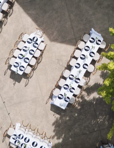 Aerial view of two long banquet tables with white tablecloths and place settings, arranged outdoors on a concrete surface with some nearby greenery.