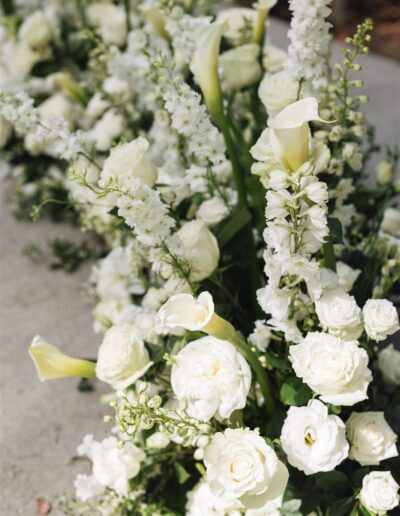 Arrangement of white flowers, including roses, calla lilies, and delphiniums, placed on a light surface.