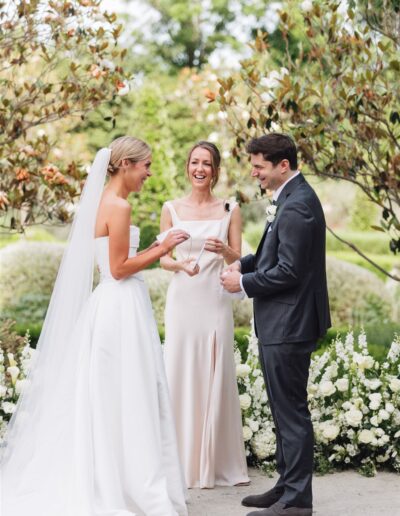 A bride and groom stand outdoors exchanging vows, smiling, with an officiant between them and white flowers in the background.