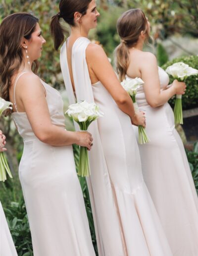 Three women in light-colored dresses stand in a row outdoors, each holding a bouquet of white flowers.