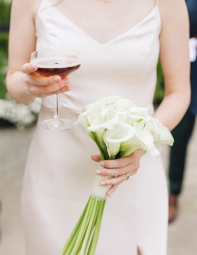 A woman in a white dress holds a bouquet of white calla lilies in one hand and a glass of red wine in the other.