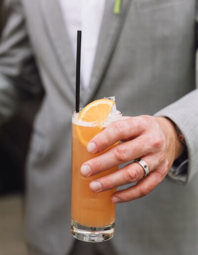 Person in a light gray suit holding a tall glass of orange-colored drink with ice, a slice of orange, and a black straw.