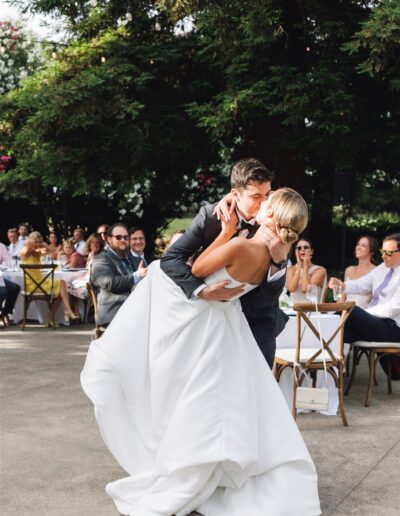 A groom in a black suit dips and kisses a bride in a white dress on an outdoor dance floor, with seated guests watching in the background.
