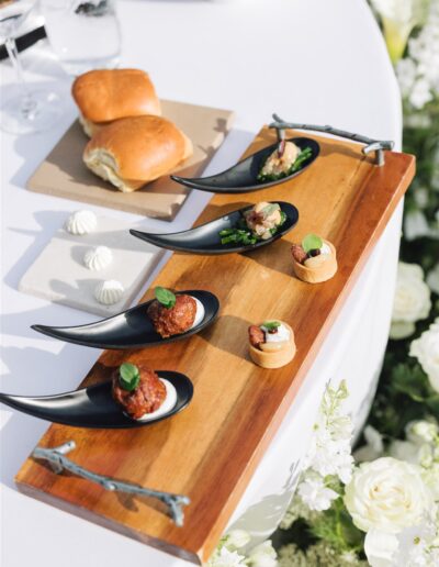 A wooden serving board with assorted bite-sized appetizers on black spoons and pastry rounds, next to bread rolls on a white table with floral decor.
