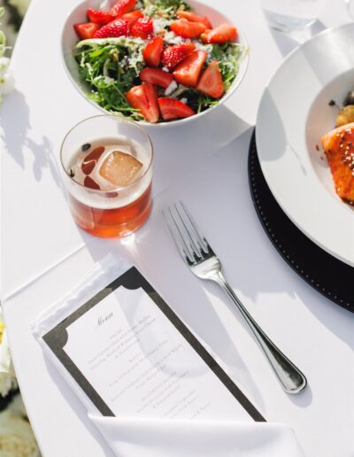 A table setting with a salad topped with strawberries, a drink with ice, a fork, a menu, and a plate of salmon on a white tablecloth.