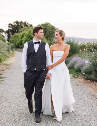 A couple in formal wedding attire walks hand in hand on a gravel path surrounded by greenery, looking at each other.