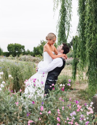 A groom lifts a bride in a white dress among wildflowers and greenery, both smiling under a weeping willow tree in an outdoor setting.