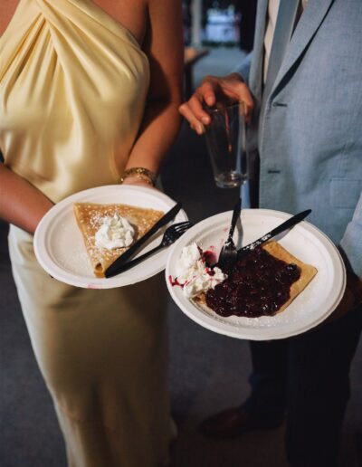 Two people holding plates with crepes; one topped with whipped cream, the other with whipped cream and berry sauce. One person holds an empty glass. Both are dressed formally.
