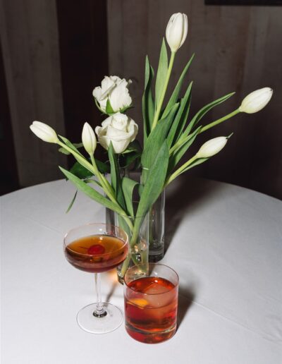 A round table with a white tablecloth holds a vase of white tulips and roses, a coupe glass with a dark cocktail and cherry, and a rocks glass with an orange-red drink and ice.