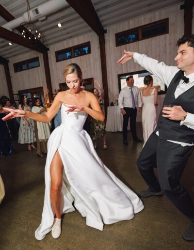 A bride in a white dress and sneakers dances energetically with guests at an indoor wedding reception.