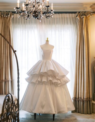 A white strapless ball gown with a tiered ruffled skirt displayed on a dress form in a room with large windows, beige curtains, and a chandelier overhead.