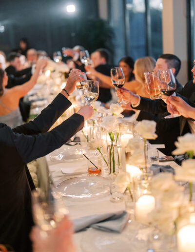 A group of people seated at a long table raise their glasses in a toast, surrounded by candles, flowers, and soft lighting.