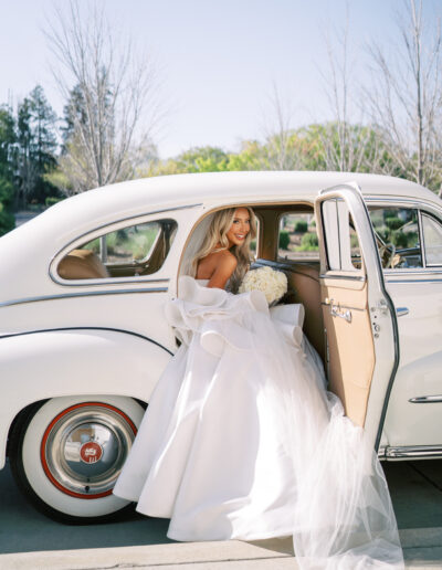 A bride in a white gown and veil steps out of a vintage white car holding a bouquet on a sunny day.