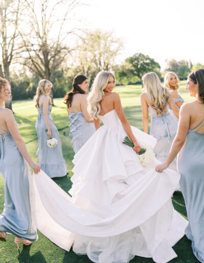 A bride in a white gown holds a bouquet and looks back while walking on grass, surrounded by bridesmaids in light blue dresses.