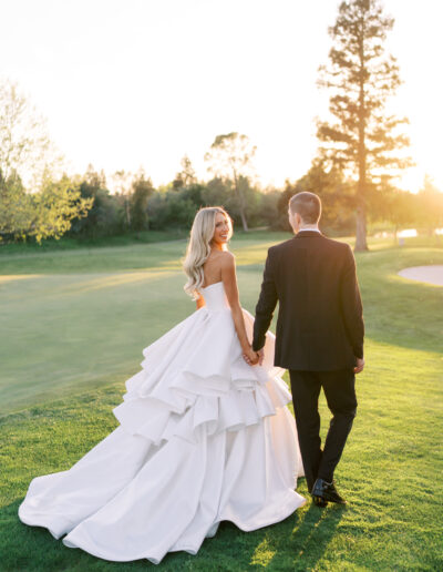 A bride in a white gown and a groom in a black suit hold hands and walk on a grassy field at sunset.