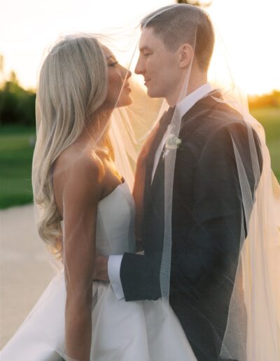 A bride and groom stand close together outdoors at sunset, wrapped in a veil, looking into each other's eyes.