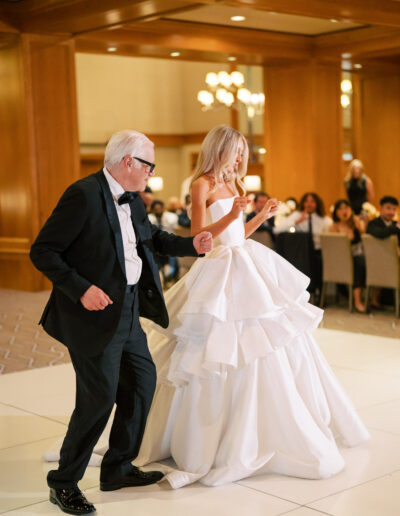 An older man in a black tuxedo and a woman in a white wedding gown dance together on a white floor at an indoor event with seated guests in the background.