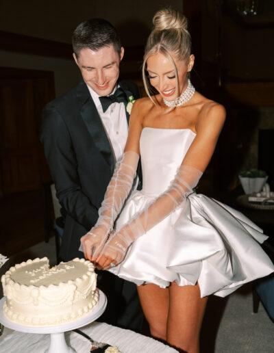 A bride and groom, both smiling, cut a white frosted cake together at their wedding reception.
