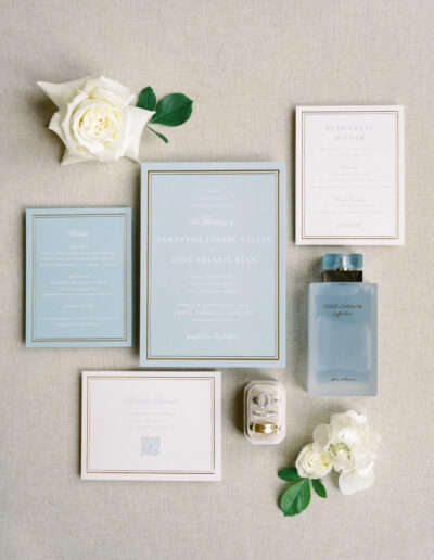 A flat lay of wedding stationery, including invitations, RSVP cards, a rehearsal dinner card, perfume, two white roses, and a small box with rings, arranged on a neutral fabric background.