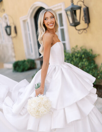 A bride in a strapless white wedding dress holds a bouquet of white roses and smiles while standing outdoors in front of a yellow building.