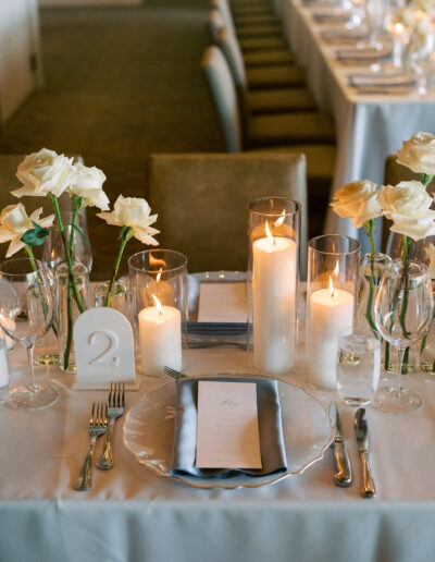 Elegant table setting with white roses, candles, glassware, silver cutlery, a translucent charger plate, and a table number "2" on a white tablecloth.