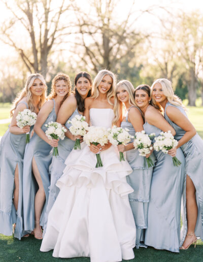 A bride in a white gown stands outdoors with six bridesmaids in light blue dresses, all holding bouquets and smiling at the camera.