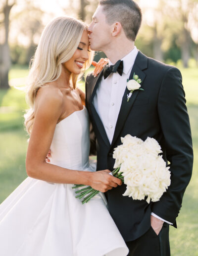 A bride in a strapless white gown holds a bouquet of white roses as a groom in a black tuxedo kisses her forehead outdoors.