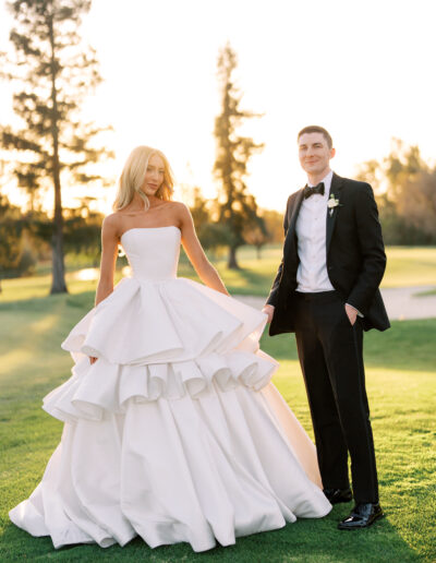 A bride in a strapless white gown with layered ruffles stands beside a groom in a black tuxedo on a grassy lawn at sunset.