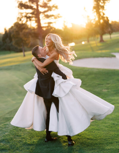 A groom lifts and spins the bride on a golf course at sunset, both smiling and dressed in formal wedding attire.