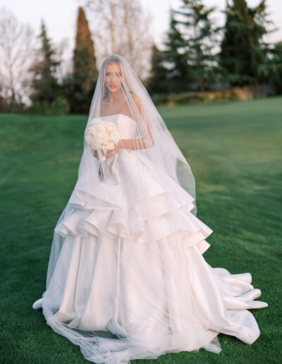 A bride in a strapless white wedding gown with a layered skirt and a long veil holds a bouquet of white flowers while standing on a grassy area outdoors.