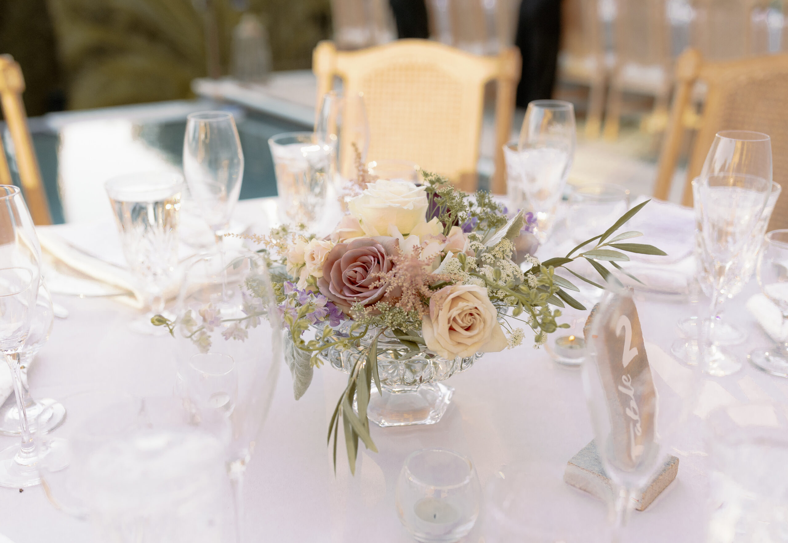 A glass vase with pastel roses and greenery serves as the centerpiece on a white-clothed table set with glassware and plates.