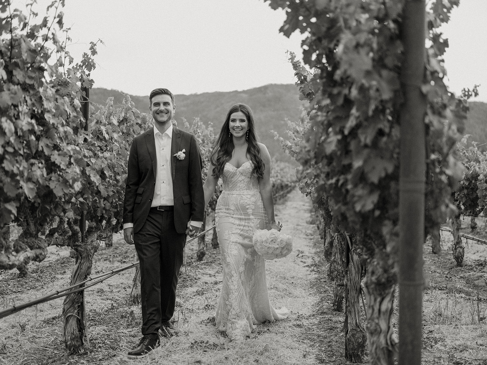 A couple dressed in wedding attire walks through a vineyard, smiling and holding hands. The background shows grapevines and distant hills.