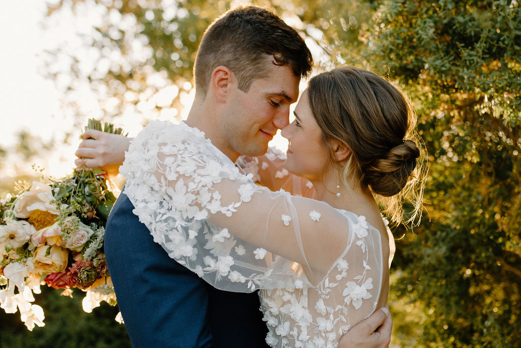 A bride and groom embrace outdoors, touching foreheads and smiling softly, with sunlight filtering through trees in the background. The bride holds a bouquet.