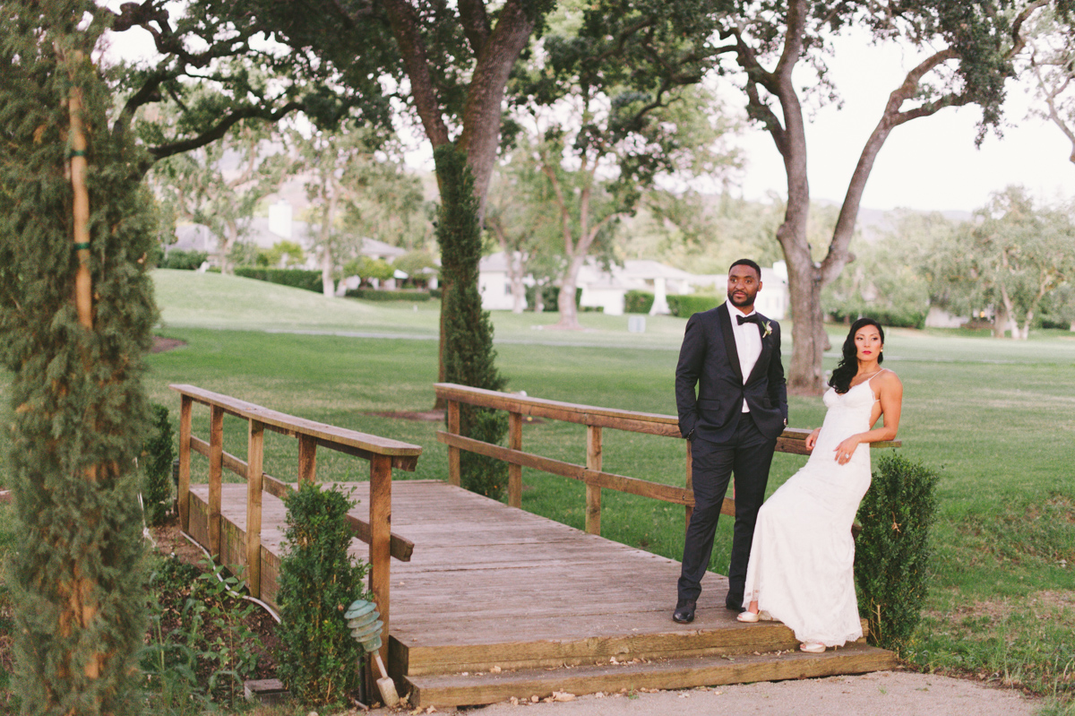 A man in a suit and a woman in a white dress stand on a wooden bridge in a park-like setting with green grass and trees.