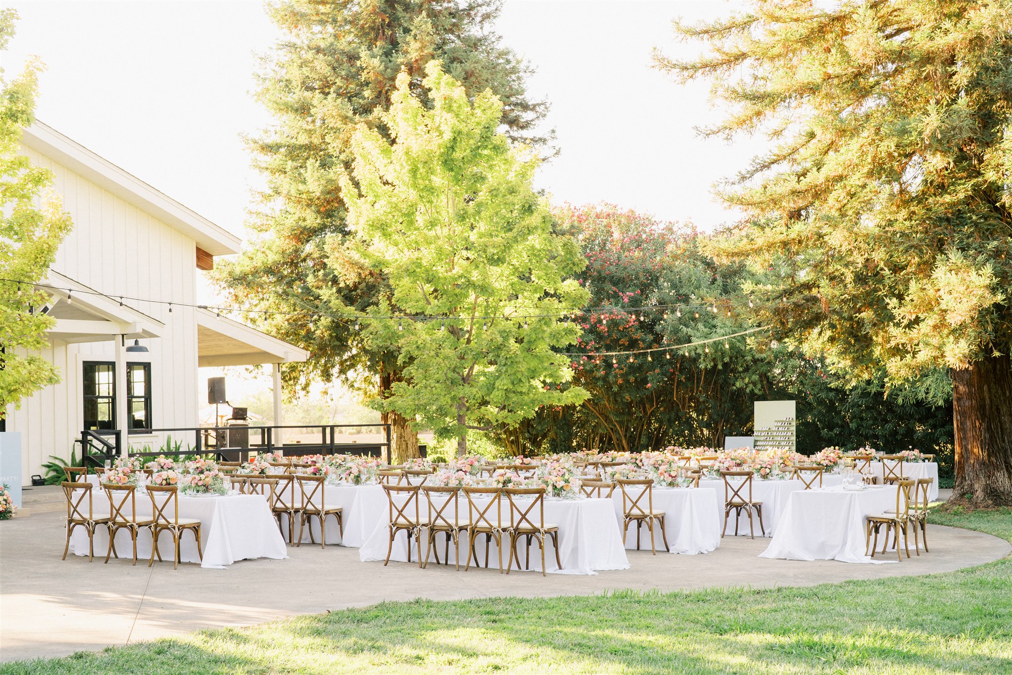 Round tables with white tablecloths and wooden chairs are arranged outdoors near a white building, surrounded by trees and greenery.