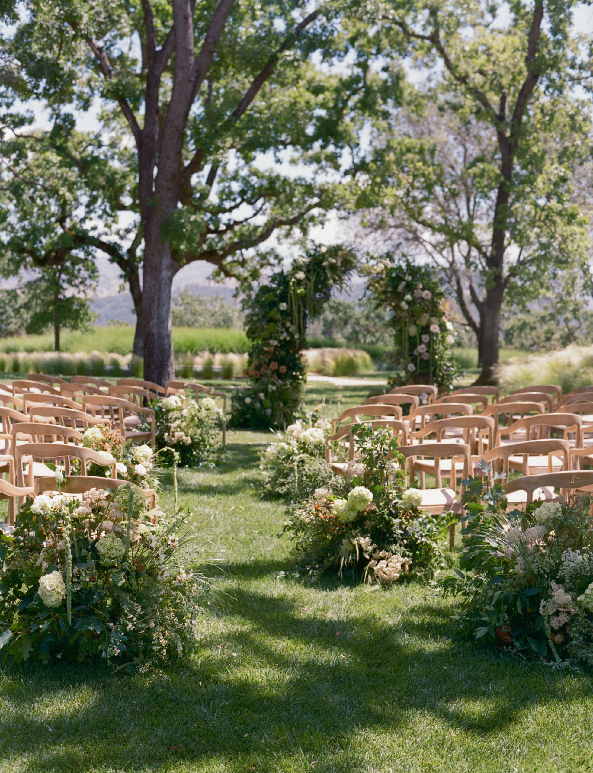 Rows of wooden chairs and floral arrangements set up outdoors on a grassy lawn, facing a decorated archway under large trees.