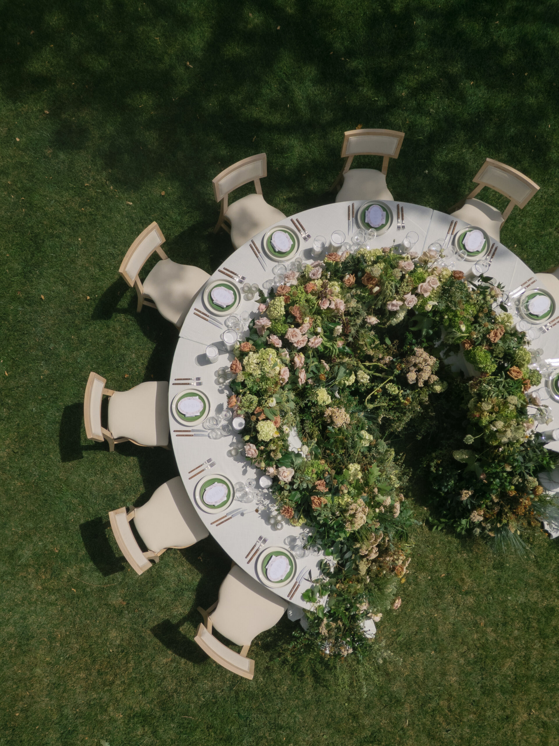 Aerial view of a round outdoor table set for a meal, decorated with abundant greenery and flowers, surrounded by chairs on a grassy lawn—an inspiring setup by Sacramento Wedding Planners.